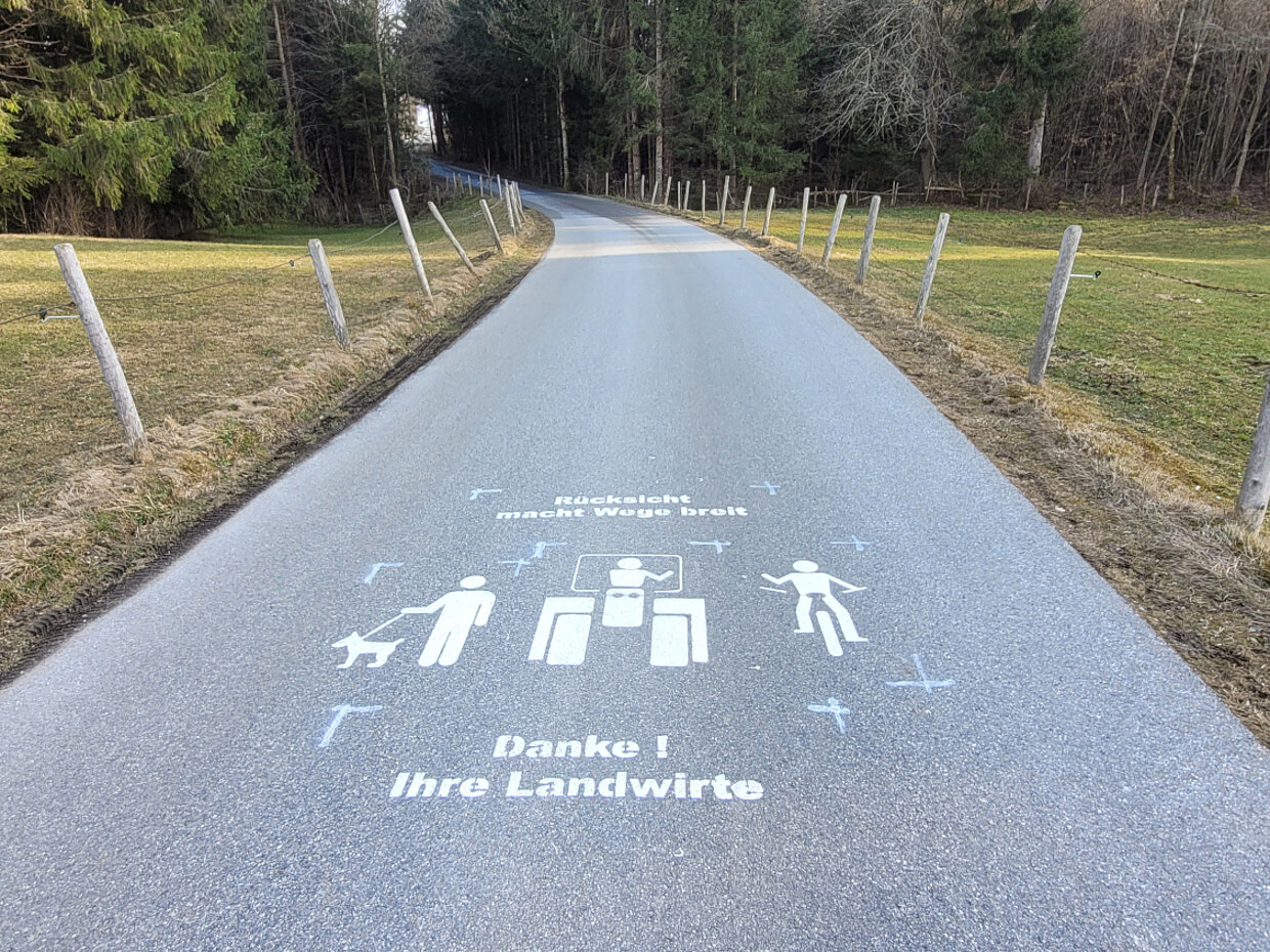 © Landkreis Ostallgäu Ein schmaler, asphaltierter Weg durch eine ländliche Umgebung, an beiden Seiten von Wiesen und Bäumen gesäumt. Auf dem Weg sind Symbole für Fußgänger, Radfahrer und einen Hund abgebildet, sowie der Text "Danke! Ihre Landwirte".
