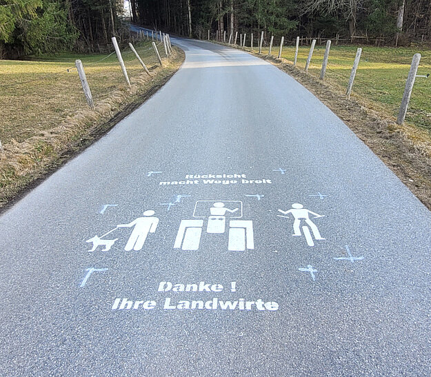 © Landkreis Ostallgäu Ein schmaler, asphaltierter Weg durch eine ländliche Umgebung, an beiden Seiten von Wiesen und Bäumen gesäumt. Auf dem Weg sind Symbole für Fußgänger, Radfahrer und einen Hund abgebildet, sowie der Text "Danke! Ihre Landwirte".