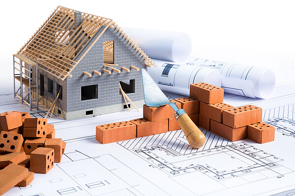 Ein Modell eines Hauses aus Holz mit einem Dach, umgeben von Bausteinen und Bauplänen. Ein Mauerwerkskelle liegt auf den Plänen. Die Szene vermittelt den Eindruck eines Bauprojekts.