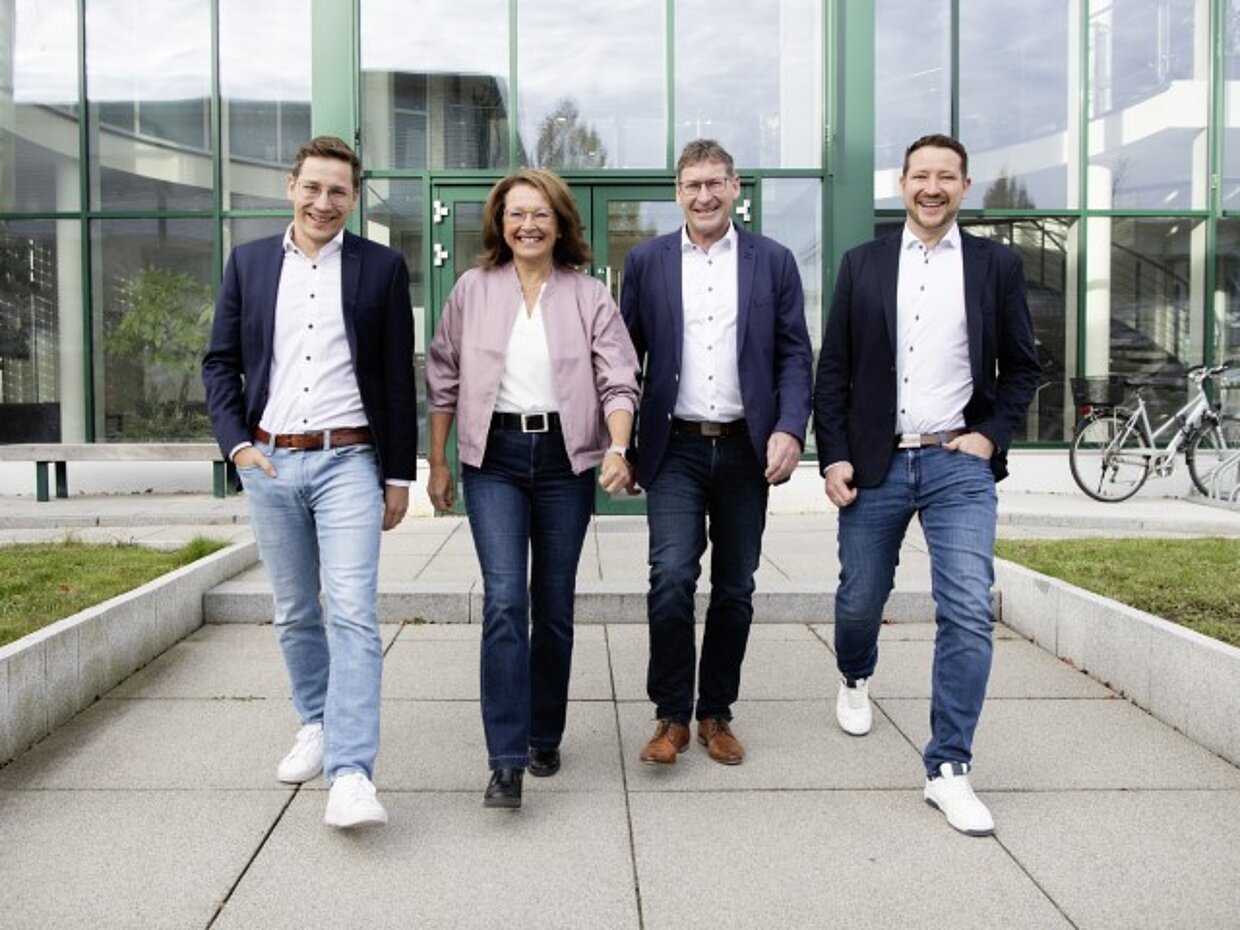© Rudolf Hörmann GmbH & Co. KG Vier Personen in formeller Kleidung gehen gemeinsam auf einem gepflasterten Weg vor einem großen Gebäude mit Glasfront. Sie lächeln und scheinen eine positive Ausstrahlung zu haben. Im Hintergrund sind Fahrräder und grüne Pflanzen sichtbar.
