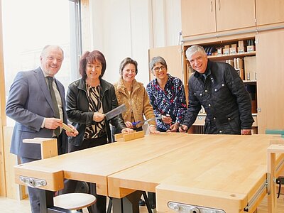 Fünf Personen stehen um einen großen Holztisch in einem hellen Raum. Sie halten Werkzeuge in der Hand und lächeln. Im Hintergrund sind Regale mit Materialien zu sehen. Das Ambiente wirkt freundlich und einladend.
