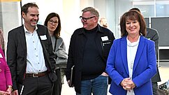 Eine Gruppe von fünf Personen steht in einer modernen Umgebung. Alle schauen lächelnd, während eine Frau mit dunklen Haaren und einem blauen Blazer im Vordergrund steht. Die anderen Personen, darunter Männer und Frauen in formeller Kleidung, wirken interessiert oder amüsiert.