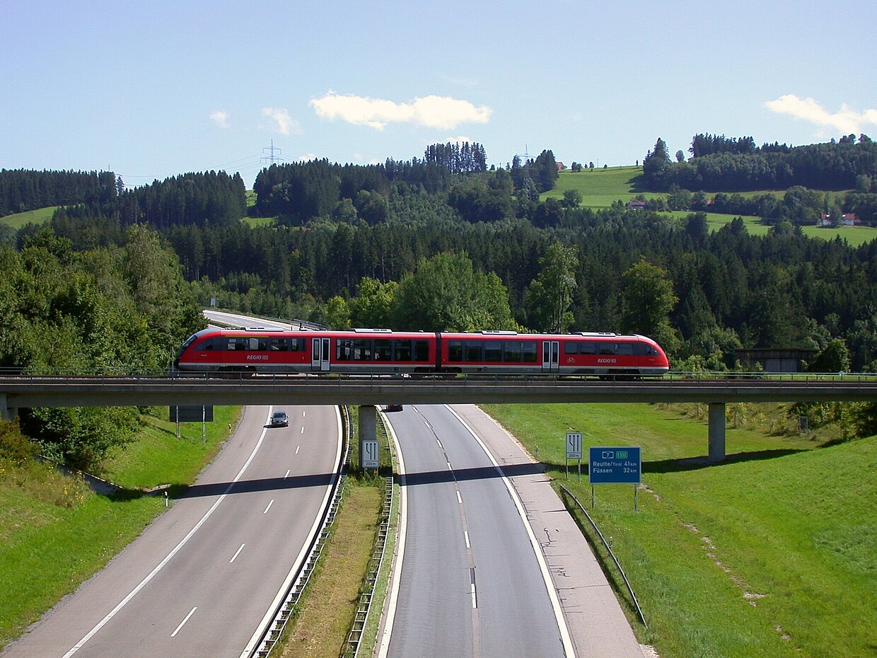 Ein roter Zug fährt auf einer Brücke, die über eine Autobahn führt. Im Hintergrund sind grüne Hügel und Wald zu sehen, unter einem klaren blauen Himmel mit wenigen Wolken.