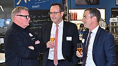 Drei Männer stehen in einem sozialen Umfeld, unterhalten sich und halten jeweils ein Glas Getränk. Der Hintergrund zeigt eine Bar mit verschiedenen Getränken und einem informellen Setting. Ein freundlicher Austausch zwischen den Männern ist deutlich erkennbar.