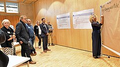 Eine Gruppe von Menschen steht in einem Raum und beobachtet eine Frau, die auf ein Flipchart schreibt. Im Hintergrund hängen zwei Informationsplakate. Die Anwesenden scheinen an einer Präsentation oder Diskussion teilzunehmen.