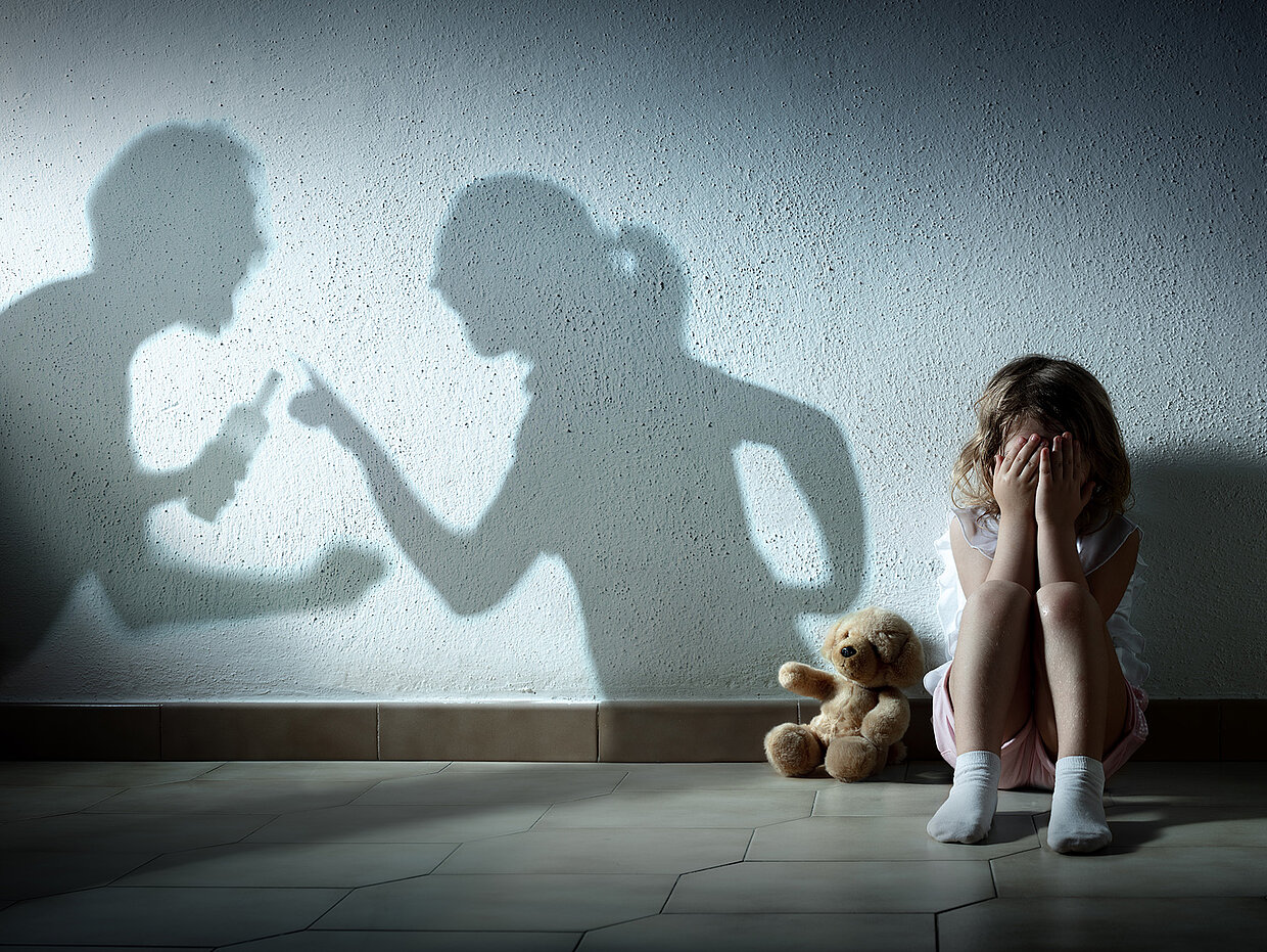 Ein kleines Mädchen sitzt mit den Händen vor dem Gesicht auf dem Boden, während Schatten von streitenden Erwachsenen an der Wand projiziert werden. Neben ihr liegt ein Stofftier. Die Szene vermittelt ein Gefühl von Angst und Unsicherheit.