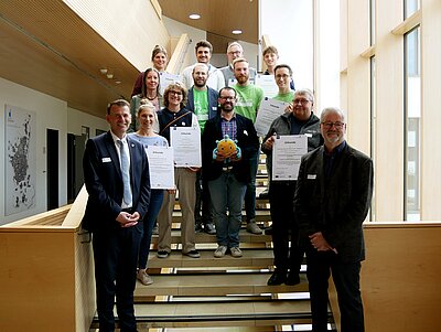 Eine Gruppe von Menschen steht auf einer Treppe in einem Gebäude. Sie halten Urkunden und einen großen, plüschigen Fisch in der Mitte der Gruppe. Hinter ihnen sind große Fenster, die Tageslicht hereinlassen. Es scheint eine feierliche Veranstaltung oder Zeremonie zu sein.