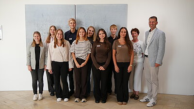 Gruppe von zwölf Personen, die lächelnd vor einer neutralen Wand stehen. Männer und Frauen unterschiedlichen Alters tragen legere Kleidung in verschiedenen Farben. Künstlerisches Hintergrundbild ist zu sehen. Die Atmosphäre ist freundlich und einladend.