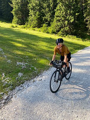 Eine Person fährt lächelnd mit einem Fahrrad auf einem Schotterweg durch eine grüne Landschaft. Von Bäumen gesäumte Wiesen sind im Hintergrund sichtbar, während die Sonne eine angenehme Atmosphäre schafft.