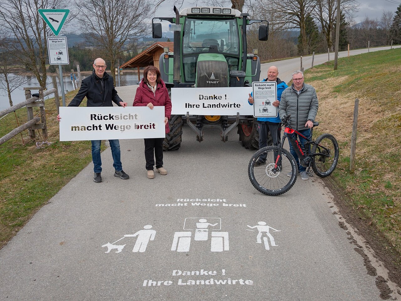 Eine Gruppe von vier Personen steht auf einem Weg. Sie halten Schilder mit der Aufschrift "Rücksicht macht Wege breit" und "Danke! Ihre Landwirte." Hinter ihnen steht ein Traktor. Auf dem Boden sind Symbole von Fußgängern, einem Hund und einem Radfahrer zu sehen.