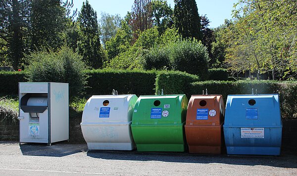 Eine Reihe von Abfallcontainern für Glasrecycling in verschiedenen Farben (weiß, grün, orange, blau) steht vor einem Hintergrund aus Bäumen und Büschen. Einige Container sind mit Hinweisen zum Recycling beschriftet.