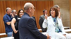 Eine Gruppe von Personen steht in einem Raum. Im Vordergrund diskutieren zwei Menschen, während andere im Hintergrund beobachten und lächeln. Der Mann trägt eine Brille und einen dunklen Anzug, die Frau einen hellen Anzug und gestikuliert. Die Atmosphäre scheint freundlich und informell zu sein.