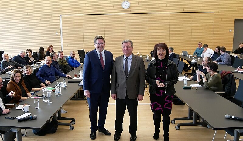 Drei Personen stehen in einem Konferenzraum, während im Hintergrund eine Gruppe von Menschen an Tischen sitzt. Die beiden Männer tragen Anzüge, die Frau ist in einem schwarzen Outfit mit bunten Akzenten gekleidet. Eine Uhr hängt an der Wand.
