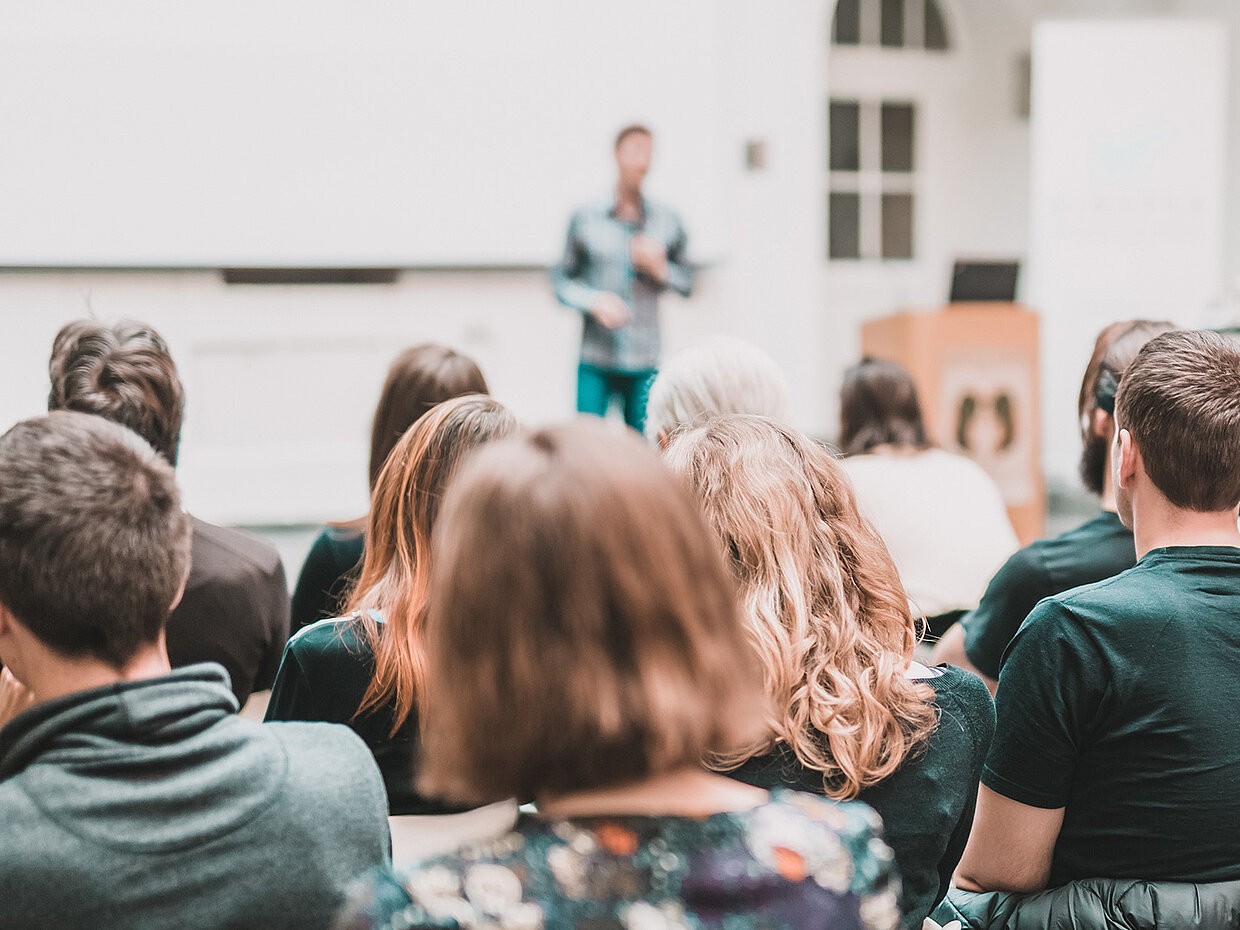 Eine Gruppe von Personen sitzt in einem Seminarraum, der Blick richtet sich auf einen Vortragenden. Der Vortragende steht im Vordergrund und spricht zum Publikum, das aus mehreren Teilnehmern besteht, die unterschiedliche Frisuren und Kleidung tragen.