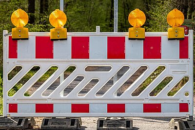 Bauzaun mit rot-weißen Streifen und gelben Blinklichtern oben. Der Zaun steht auf einem Weg, um eine Baustelle oder Straßensperrung anzuzeigen.