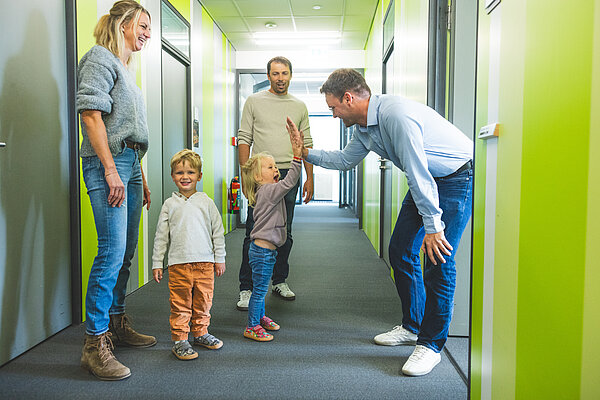 Eine Familie steht in einem hellen Flur. Zwei kleine Kinder versuchen, ein High-Five mit einem erwachsenen Mann zu geben. Eine Frau und ein weiterer Mann beobachten die Szene lächelnd. Der Flur hat grüne Wände und ist gut beleuchtet.