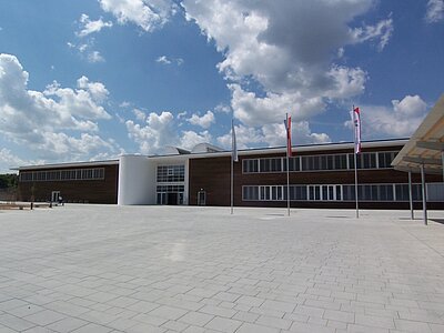 Bildbeschreibung: Ein modernes Gebäude mit großen Fenstern und einer Holzfassade, umgeben von einem gepflasterten Platz. Der Himmel ist klar mit einigen Wolken, und es stehen mehrere Fahnen vor dem Gebäude.
