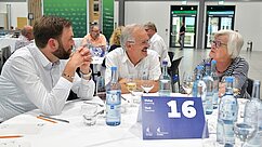 Drei Personen sitzen an einem Tisch und unterhalten sich. Eine Frau mit Brille und grau-weißem Haar hört zu, während ein Mann mit Bart und ein weiterer Mann mit Brille aktiv sprechen. Auf dem Tisch steht eine Flasche Wasser und Gläser, sowie eine Tischnummer 16.