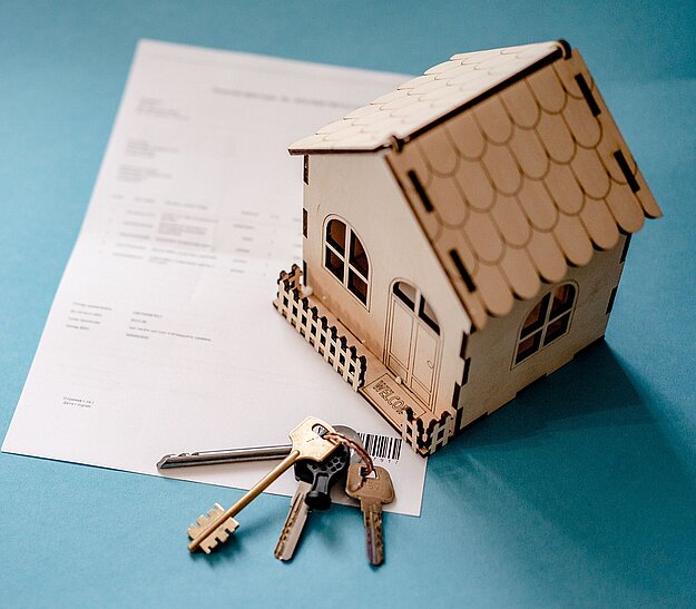 Eine hölzerne Modellhaus steht neben einem Vertrag und einem Schlüsselbund auf einer blauen Hintergrundfläche. Der Vertrag zeigt möglicherweise Informationen zu Immobilien. Die Schlüssel symbolisieren den Besitz oder den Zugang zu einem neuen Zuhause.