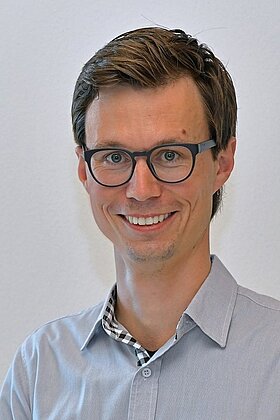 Ein junger Mann mit braunen Haaren und einer Brille lächelt freundlich. Er trägt ein hellblaues Hemd mit einem Karomuster im Kragen. Der Hintergrund ist schlicht und hell gestaltet.
