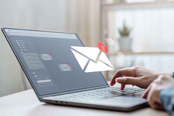 Eine Person tippt auf der Tastatur eines Laptops, während eine animierte E-Mail-Icon mit einer Benachrichtigung über eine neue Nachricht angezeigt wird. Im Hintergrund sind verschwommene Fenster und Pflanzen zu sehen.