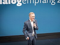 Ein Mann mit Brille und Anzug steht auf einer Bühne und spricht in ein Mikrofon. Im Hintergrund ist eine große blaue Wand mit dem Text „Dialogempfang 2023“ und einem Logo der Wirtschaft Ostallgäu zu sehen.