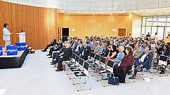 Eine Person steht an einem Rednerpult vor einem Publikum in einem modernen Innenraum. Viele Zuhörer sitzen in Reihen, gut gekleidet, und blicken auf die Rednerin. Der Raum hat große Fenster und eine Holzverkleidung.