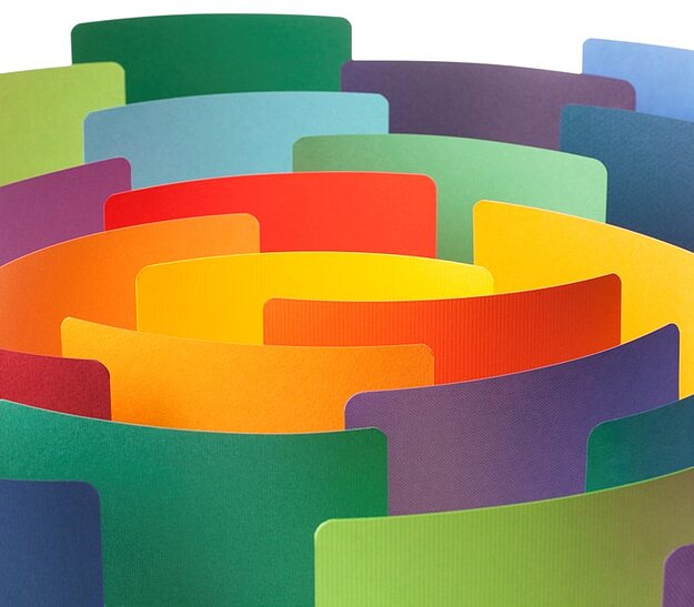 Bunte, geschichtete Papiere in spiralförmiger Anordnung, die eine ansprechende visuelle Komposition bilden. Die Farben reichen von Rot über Gelb, Grün bis Blau und Violett. Die Textur der Papiere ist glatt und wirkt dynamisch.