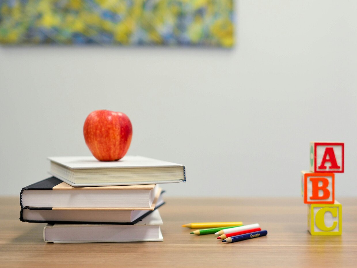 Ein Stapel Bücher steht auf einem Tisch, mit einem roten Apfel obenauf. Neben den Büchern befinden sich einige Buntstifte und ein Block mit bunten Bauklötzen, die die Buchstaben A, B und C zeigen. Im Hintergrund ist ein abstraktes Gemälde sichtbar.