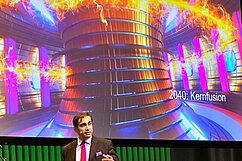 Ein Mann in Anzug und Krawatte steht vor einer Projektionsleinwand, die ein futuristisches Kernfusionsgerät zeigt. Das Bild zeigt leuchtende Farben und dynamische Energie, während die Jahreszahl 2040 neben der Grafik steht. Der Mann spricht über die Zukunft der Kernfusion.