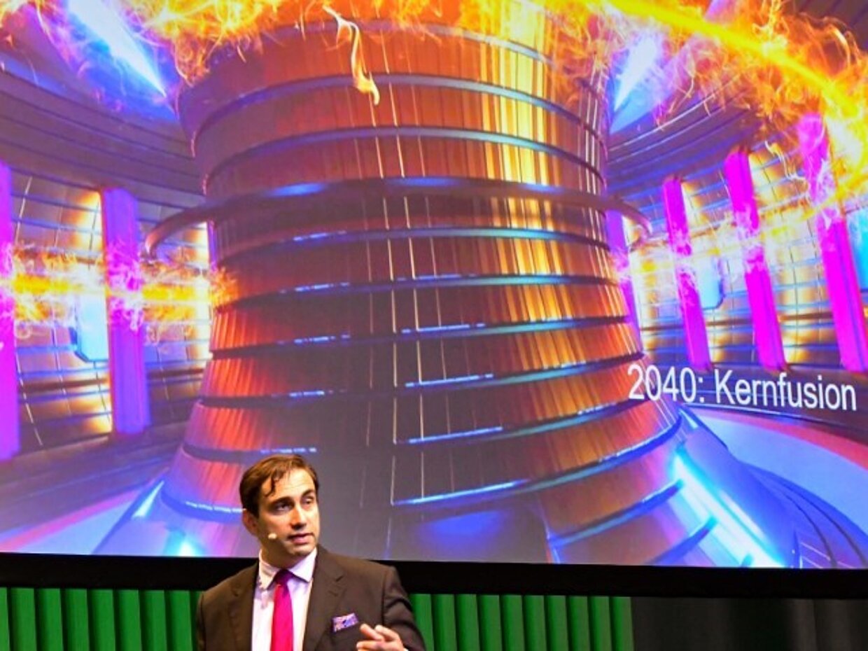 Ein Mann in Anzug und Krawatte steht vor einer Projektionsleinwand, die ein futuristisches Kernfusionsgerät zeigt. Das Bild zeigt leuchtende Farben und dynamische Energie, während die Jahreszahl 2040 neben der Grafik steht. Der Mann spricht über die Zukunft der Kernfusion.