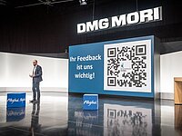 Ein Vortragender steht auf einer Bühne vor einer großen Leinwand mit einem QR-Code und dem Text „Ihr Feedback ist uns wichtig!“. Im Hintergrund sieht man das Logo von DMG MORI und einige Podeste.