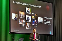 Ein Referent steht auf einer Bühne vor einer Präsentation mit dem Titel "Kann man Zukunft vorhersagen?" und zeigt Bilder von verschiedenen Experten. Darunter sind Namen von internationalen Führungskräften aus Technologie und Innovation aufgelistet.