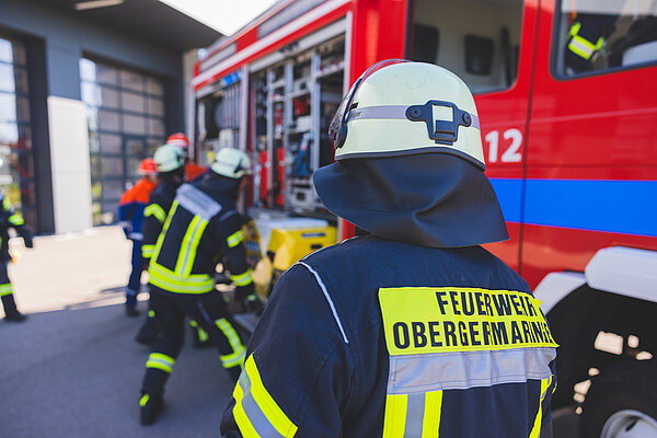 Feuerwehrleute in Schutzkleidung arbeiten an einem Feuerwehrfahrzeug. Im Vordergrund steht ein Feuerwehrmann mit einer Jacke, auf der "FEUERWEHR OBERGEM." steht. Im Hintergrund sind weitere Feuerwehrleute sichtbar, die Ausrüstung entladen.
