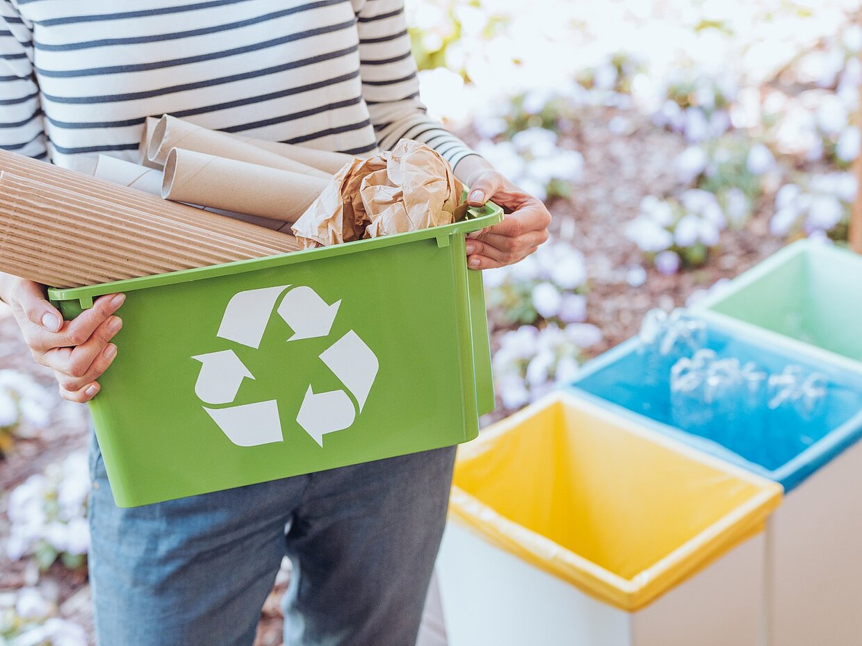 Eine Person hält einen grünen Recyclingbehälter, gefüllt mit Papier und Kartonagen, vor drei Abfallbehältern in verschiedenen Farben. Der grüne Behälter zeigt ein Recycling-Symbol. Im Hintergrund sind Blumen sichtbar.