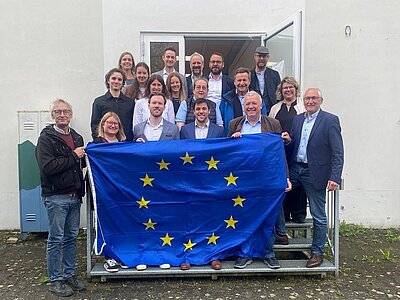 Eine Gruppe von 19 Menschen steht vor einem Gebäude, einige halten eine EU-Flagge. Sie lächeln und wirken fröhlich, während sie gemeinsam posieren. Das Bild zeigt ein Gefühl von Gemeinschaft und Zugehörigkeit zur Europäischen Union.