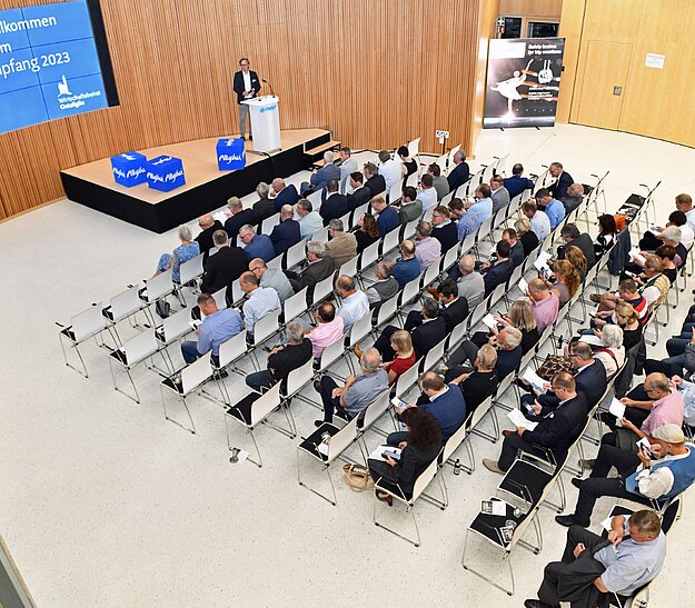 Eine große Gruppe von Menschen sitzt in einem modernen Raum. Auf der Bühne steht ein Redner, der eine Präsentation hält. An einer Wand hängt ein Bildschirm mit der Begrüßung „Herzlich Willkommen Zum Dialoganfang 2023“. Die Anordnung ist formal und die Atmosphäre scheint geschäftlich.