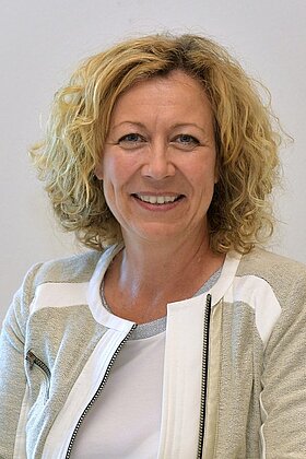 Porträt einer lächelnden Frau mit lockigem, blonden Haar. Sie trägt eine helle Jacke und sitzt vor einem einfarbigen Hintergrund. Der Ausdruck vermittelt Freundlichkeit und Offenheit.