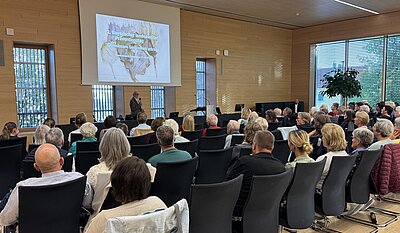 Ein Vortrag in einem modernen Raum mit großen Fenstern. Eine Gruppe von Menschen sitzt in Stühlen und schaut auf eine Präsentation, die eine Karte zeigt. Der Redner steht vor der Leinwand und erklärt etwas.