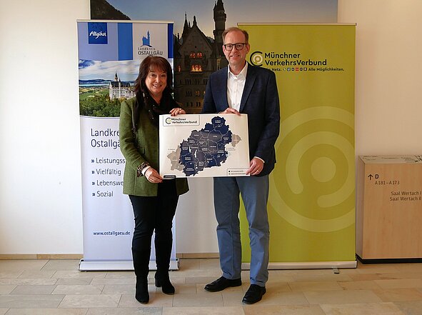 Zwei Personen stehen in einem Raum und halten eine Karte des Münchner Verkehrsverbunds. Im Hintergrund sind Banner des Landkreises Ostallgäu und des Münchner Verkehrsverbunds sichtbar. Die Person links trägt eine grüne Jacke, die rechts eine dunkle Jacke und Brille.