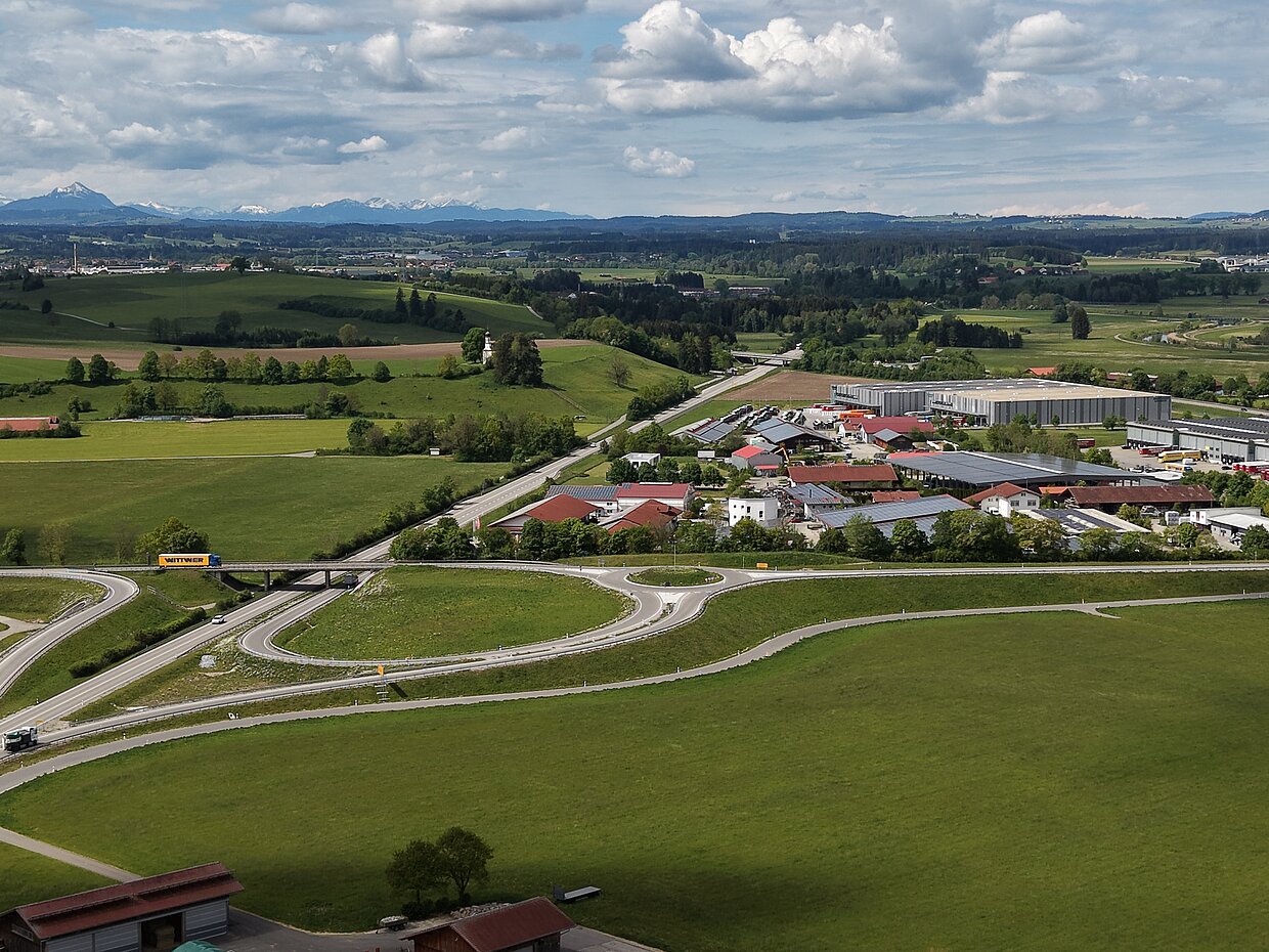 Landschaft mit Wiesen und Feldern unter einem bewölkten Himmel. Im Vordergrund sieht man eine Straßenabzweigung, wo Fahrzeuge fahren. Im Hintergrund erstrecken sich Wohnhäuser und eine Industrieanlage, während in der Ferne Berge sichtbar sind.