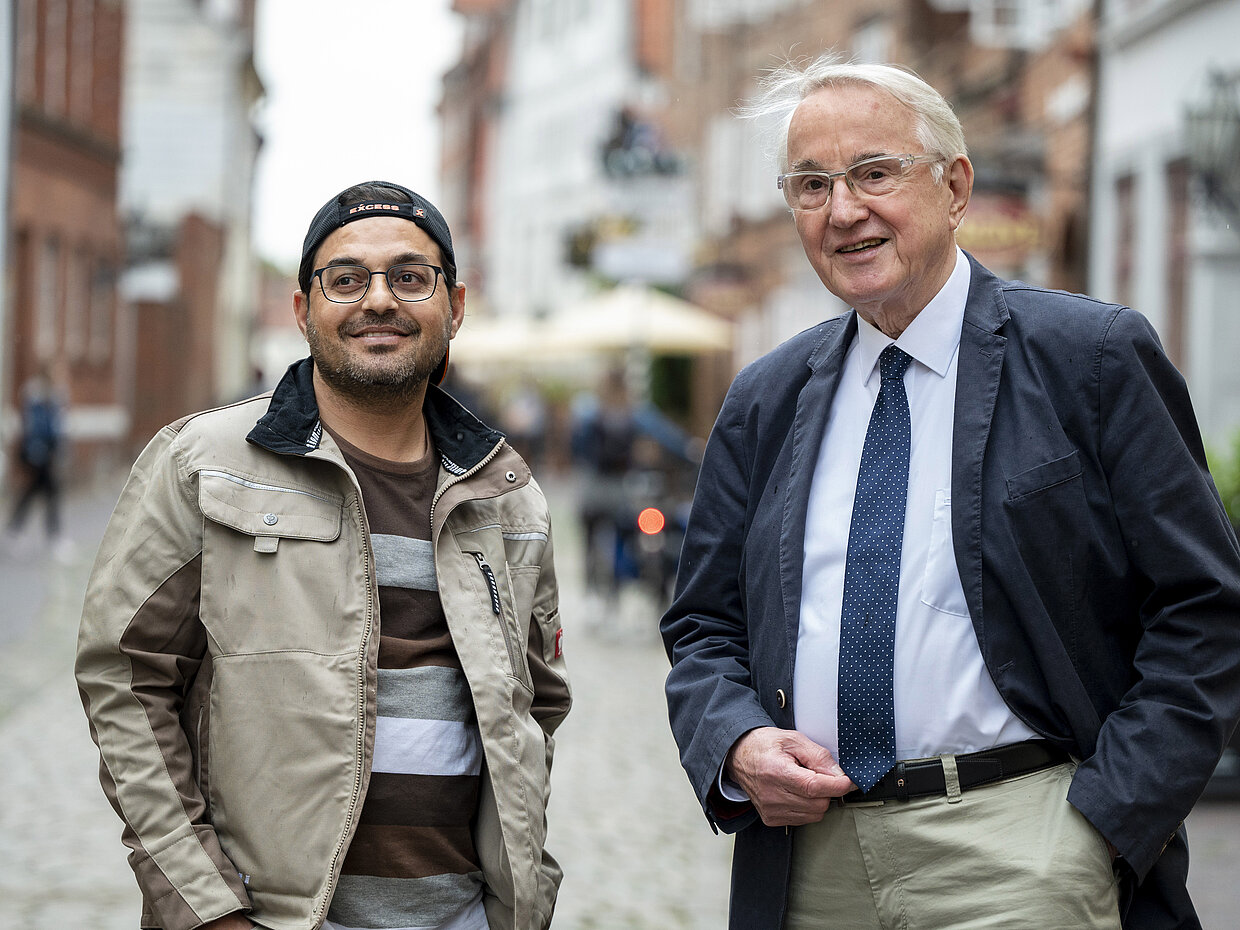 Zwei Männer stehen auf einer historischen Straße. Der junge Mann trägt eine lässige Jacke und eine Baseballmütze, während der ältere Mann Anzug und Krawatte trägt. Beide lächeln in die Kamera. Im Hintergrund sind Gebäude und Passanten zu sehen.