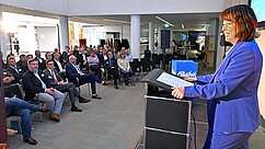 Landrätin Maria Rita Zinnecker steht hinter einem Rednerpult und spricht zu einem Publikum in einem modernen Raum. Das Publikum sitzt in Stuhlreihen und schaut aufmerksam auf die Rednerin, die in einem blauen Blazer gekleidet ist. An der Wand ist eine Präsentation sichtbar.