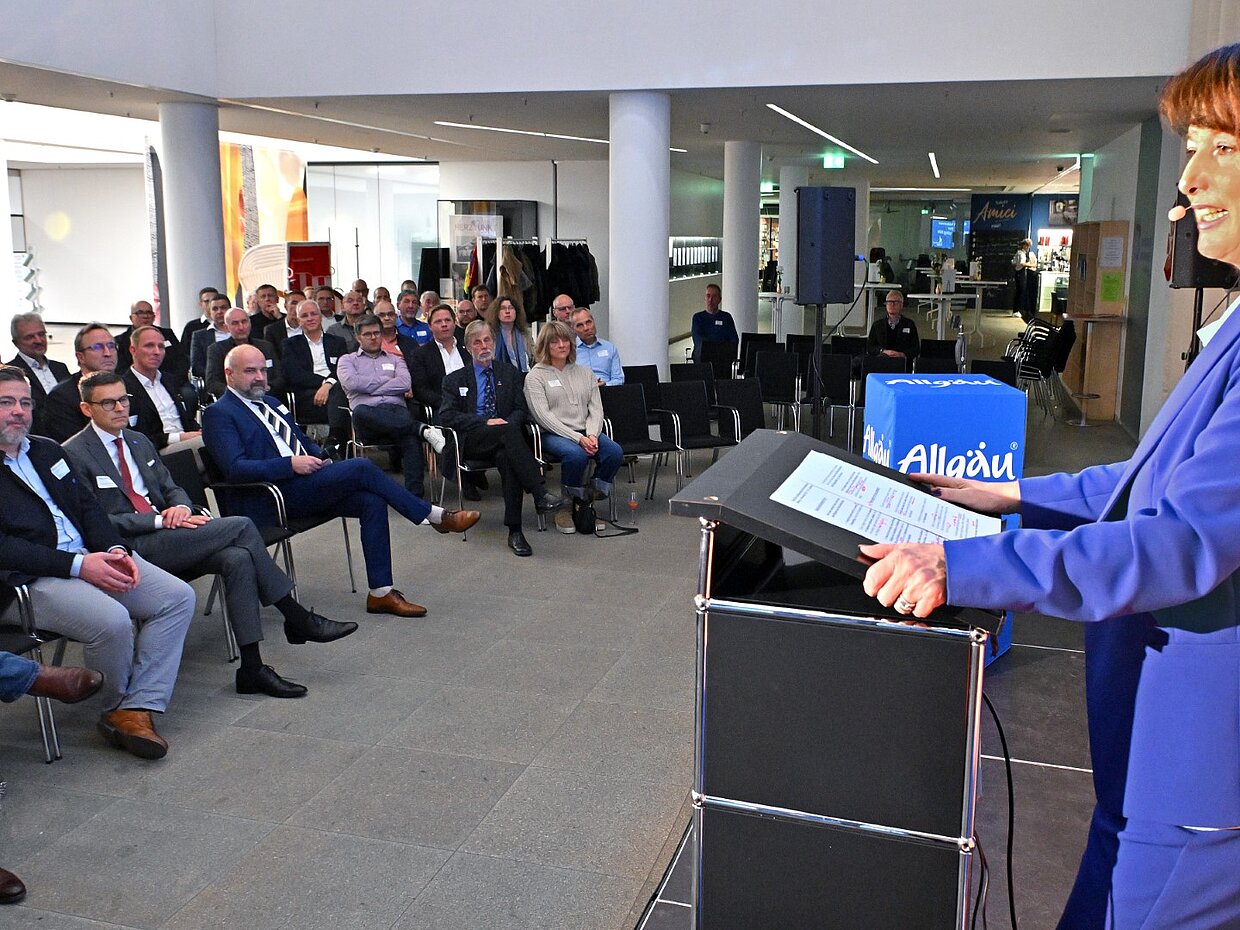 Landrätin Maria Rita Zinnecker steht hinter einem Rednerpult und spricht zu einem Publikum in einem modernen Raum. Das Publikum sitzt in Stuhlreihen und schaut aufmerksam auf die Rednerin, die in einem blauen Blazer gekleidet ist. An der Wand ist eine Präsentation sichtbar.