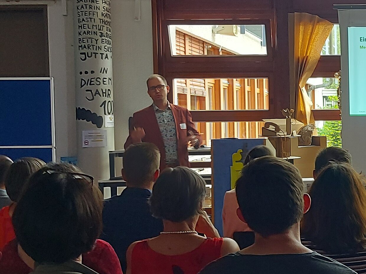 Ein Referent steht vor einem Publikum in einem hellen Raum. Die Anwesenden sitzen auf Stühlen und schauen aufmerksam zu. Im Hintergrund sind große Fenster mit Tageslicht und eine Präsentation auf einem Bildschirm sichtbar. Der Referent trägt einen orangefarbenen Anzug und erklärt etwas.