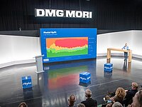 Ein Raum mit einer Präsentation. Auf der Bühne steht ein Mann vor einem großen Bildschirm, der ein Diagramm zeigt, das verschiedene Farben zur Darstellung von Daten nutzt. Im Publikum sind mehrere Zuschauer zu sehen, einige auf Stühlen. Im Hintergrund steht das Logo "DMG MORI".