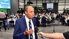Ein Mann in einem blauen Anzug und einer Krawatte spricht in ein Mikrofon. Im Hintergrund sind eine Gruppe von Menschen und landwirtschaftliche Maschinen zu sehen. Der Raum ist für eine Veranstaltung hergerichtet, mit Tischen und Stühlen.