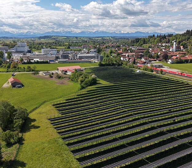 Luftaufnahme einer ländlichen Landschaft mit einem Solarfeld im Vordergrund, umgeben von grünen Wiesen und Bäumen. Im Hintergrund sind Gebäude und eine Kleinstadt zu sehen, während sich die Berge im Hintergrund abzeichnen. Ein Zug fährt entlang der rechten Bildseite.