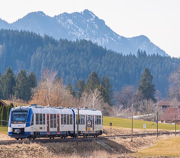 Ein moderner Zug fährt entlang einer landschaftlichen Strecke, umgeben von Bergen und dichten Wäldern. Im Hintergrund sind klare blaue Himmel und eine hügelige Landschaft zu sehen. Der Zug ist mit Fenstern ausgestattet und hat ein weiß-blaues Design.