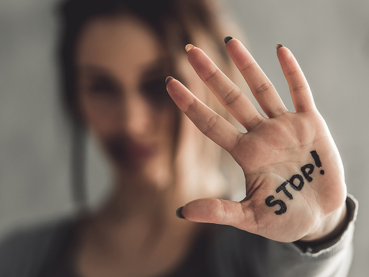 Eine unscharfe Frau hält ihre Hand mit der Aufschrift „STOP!“ nach vorne. Die Geste vermittelt eine klare Aufforderung zur Unterbrechung oder Beendigung einer Handlung. Der Fokus liegt auf der Hand und der Botschaft, während der Hintergrund verschwommen ist.